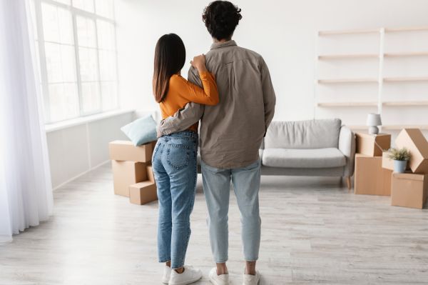 A couple stands arm in arm in a bright, unfurnished room with moving boxes, looking toward a sofa and empty shelves—ready to settle into their new space with confidence knowing effective pest control will keep their home comfortable.