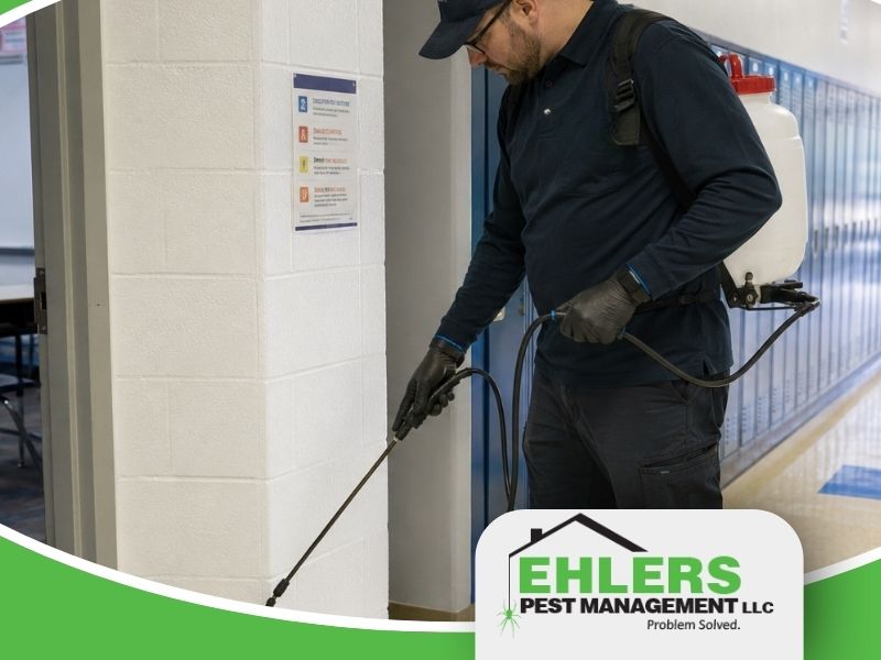 A pest control worker in uniform sprays near a wall in a hallway with blue lockers, holding a sprayer connected to a backpack tank. The Ehlers Pest Management LLC logo appears in the bottom right corner, highlighting their professional pest solutions.