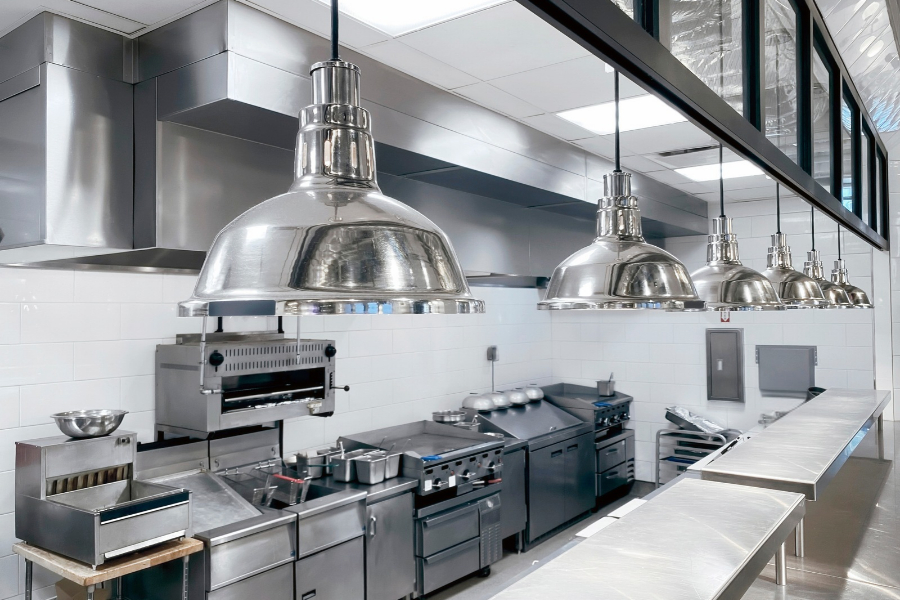 A modern commercial kitchen with stainless steel appliances, metal pendant lights, white tile walls, ovens, stoves, and various cooking equipment arranged neatly.