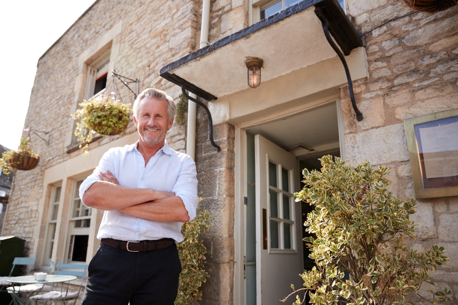 A man with gray hair stands smiling with arms crossed outside a rustic stone building, near an open door and potted plants—perfect for showcasing expert rental property pest control in Milwaukee.