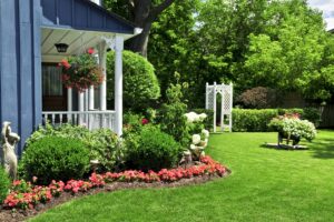 A front porch with a blue exterior and white railings overlooks a garden with blooming pink flowers, shrubs, and a white trellis in a green lawn.