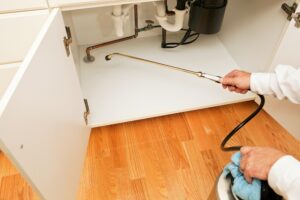 A person uses a long metal spray wand to apply pest control treatment inside an open kitchen cabinet under a sink.