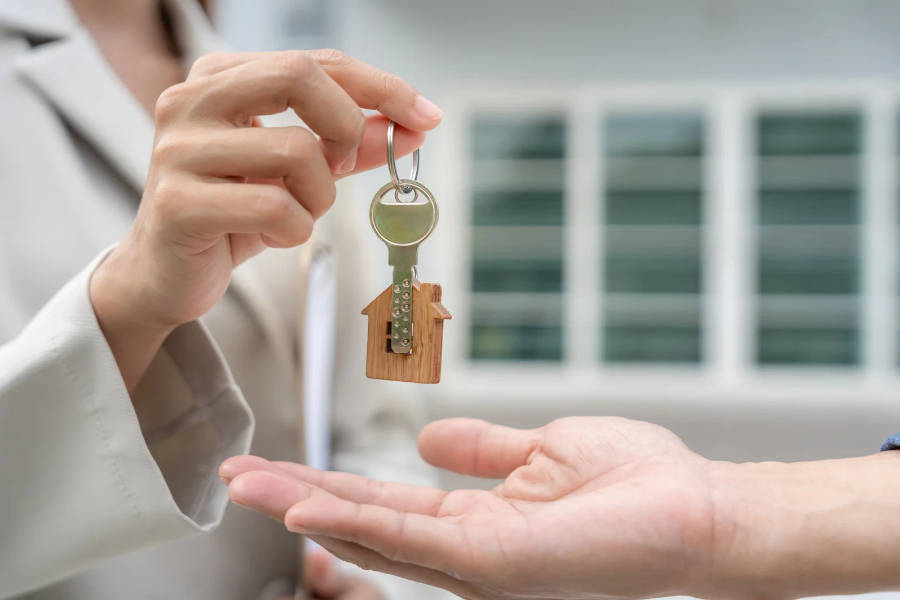 A person hands a key with a house-shaped keychain to another, symbolizing buying, selling, or renting a home—perfect for rental property pest control Milwaukee. The background is blurred with windows visible.